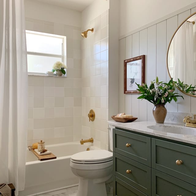 Bright bathroom with white tiles and gold fixtures. A round mirror hangs above a green vanity with flowers. Cozy, inviting, and elegant atmosphere.