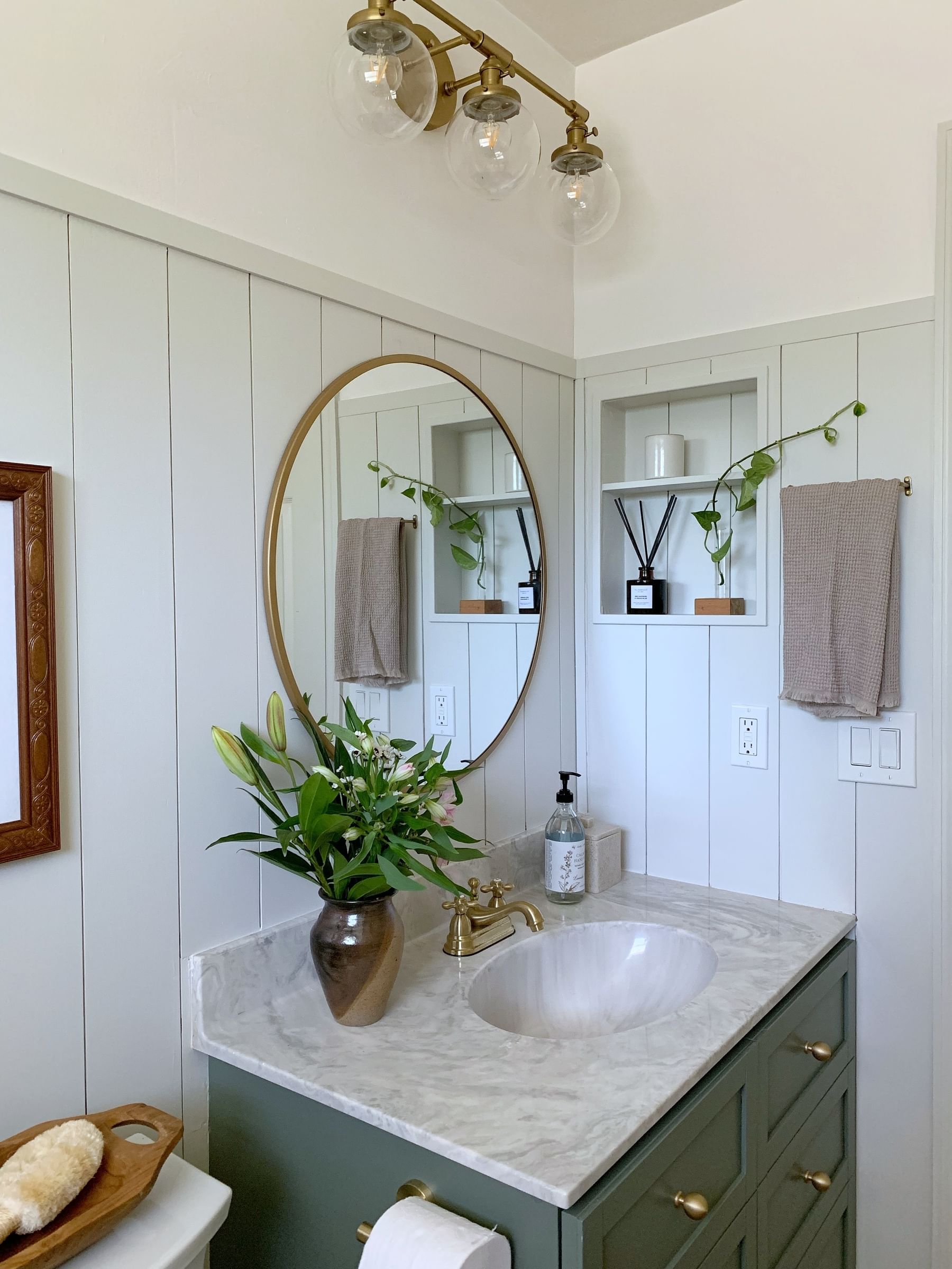 A cozy bathroom features a marble countertop with a green vanity, gold accents, a round mirror, vase of lilies, and overhead globe lights, exuding elegance.