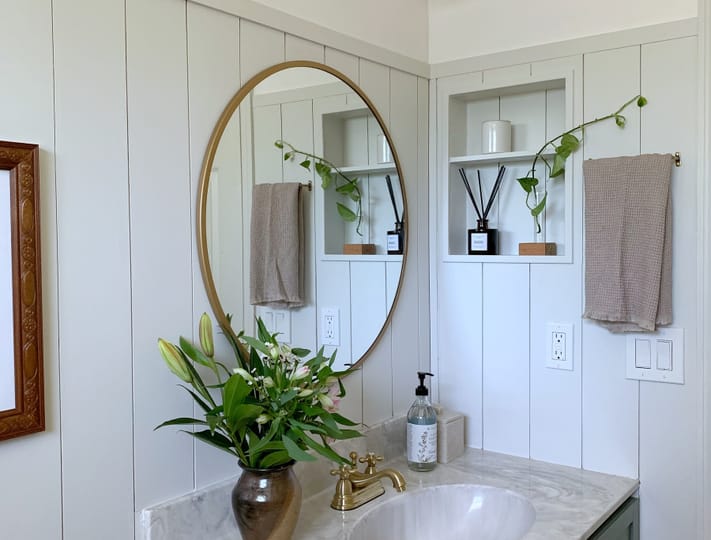 A cozy bathroom features a marble countertop with a green vanity, gold accents, a round mirror, vase of lilies, and overhead globe lights, exuding elegance.