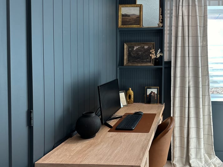 A cozy home office with a wooden desk, computer, and leather chair. Blue panel walls, shelves with art, and a window with checked curtains add warmth.