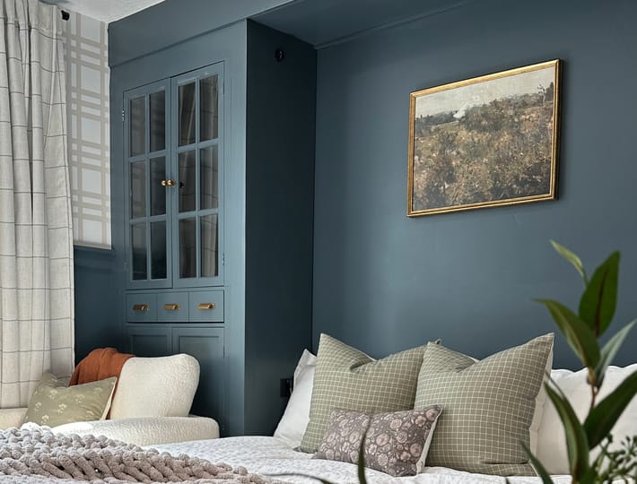 Cozy bedroom with teal walls, a white bed adorned with textured pillows and a thick knit gray blanket, a potted plant, and a landscape painting.