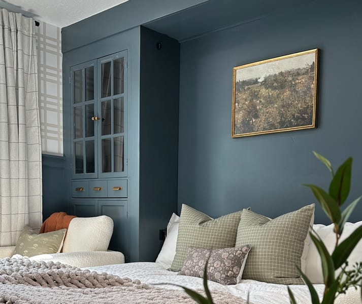 Cozy bedroom with teal walls, a white bed adorned with textured pillows and a thick knit gray blanket, a potted plant, and a landscape painting.