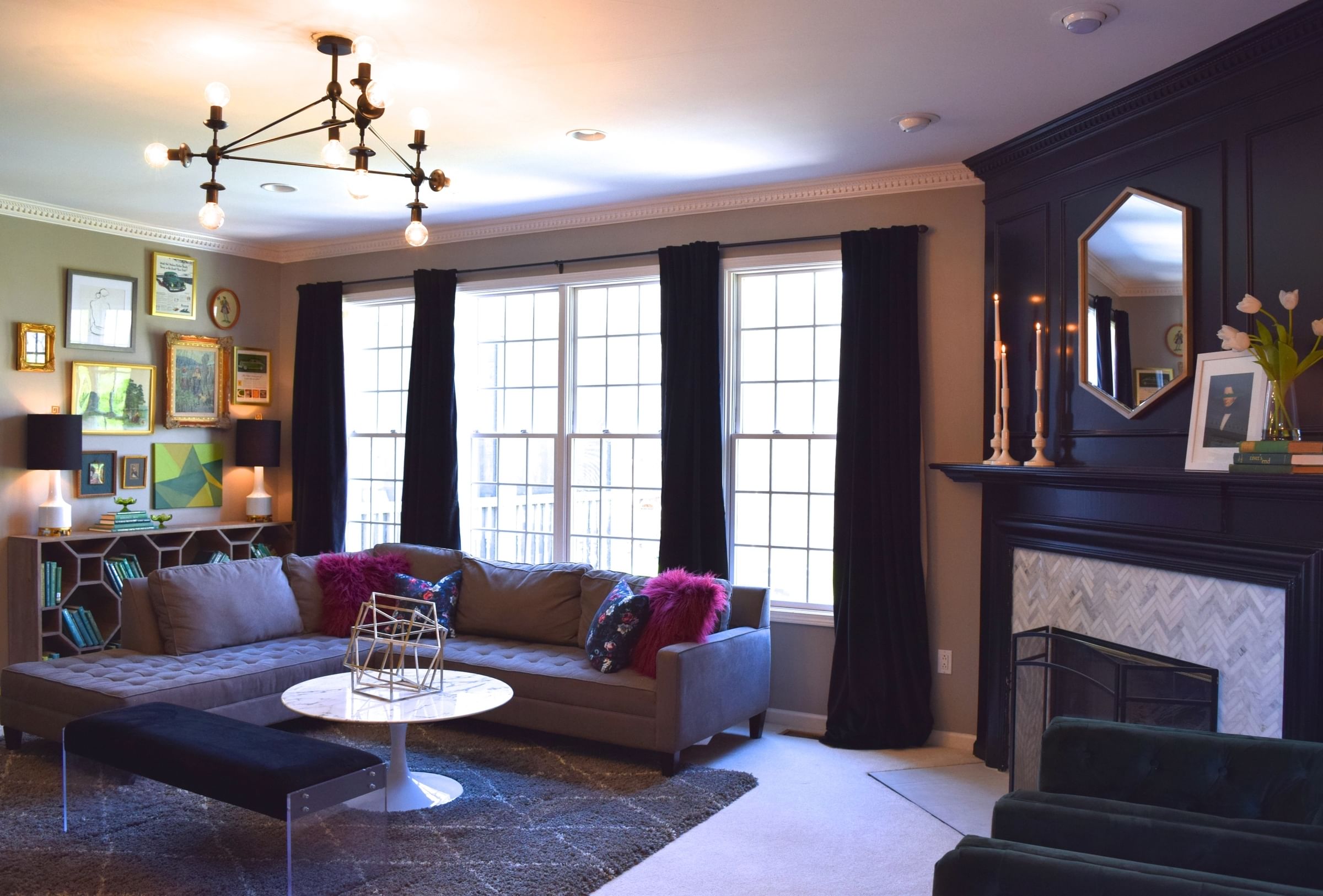 A modern living room featuring a gray sectional sofa with colorful pillows, a geometric coffee table, elegant wall art, black velvet curtains, and a stylish chandelier. The space conveys a cozy and sophisticated ambiance.