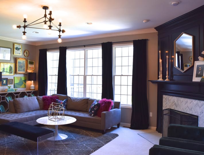 A modern living room featuring a gray sectional sofa with colorful pillows, a geometric coffee table, elegant wall art, black velvet curtains, and a stylish chandelier. The space conveys a cozy and sophisticated ambiance.