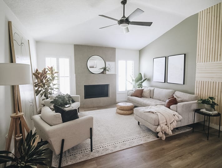 Modern living room with a beige sectional sofa, textured rug, and potted plants. A round mirror hangs above the fireplace, creating a cozy atmosphere.