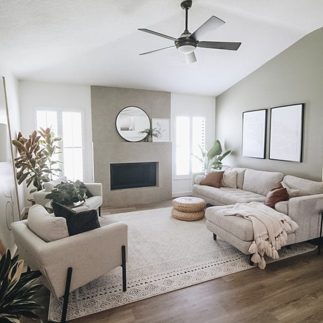 Modern living room with a beige sectional sofa, textured rug, and potted plants. A round mirror hangs above the fireplace, creating a cozy atmosphere.