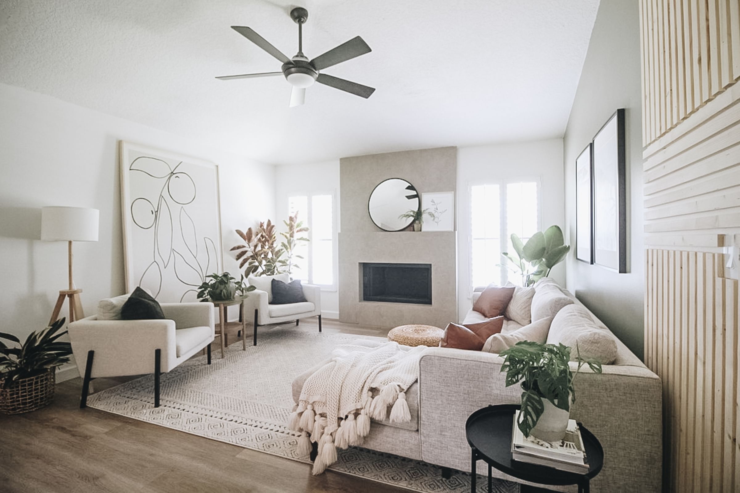 Bright, modern living room with neutral tones. Features a large beige sectional with throw pillows, abstract wall art, plants, and a cozy fireplace.