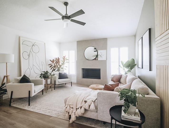 Bright, modern living room with neutral tones. Features a large beige sectional with throw pillows, abstract wall art, plants, and a cozy fireplace.
