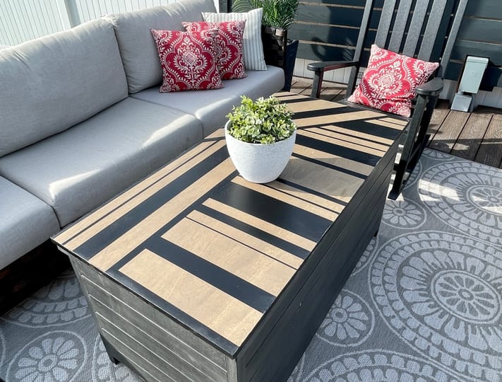Outdoor patio with a gray couch, red patterned pillows, and a black rocking chair. A striped table with a potted plant sits on a circular patterned rug.
