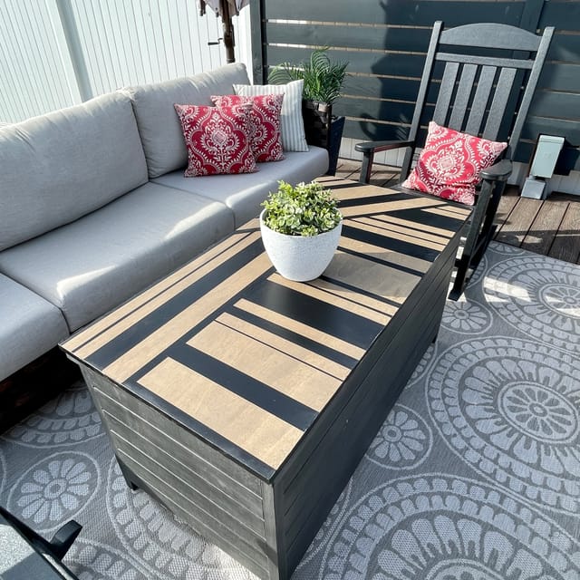 Outdoor patio with a gray couch, red patterned pillows, and a black rocking chair. A striped table with a potted plant sits on a circular patterned rug.