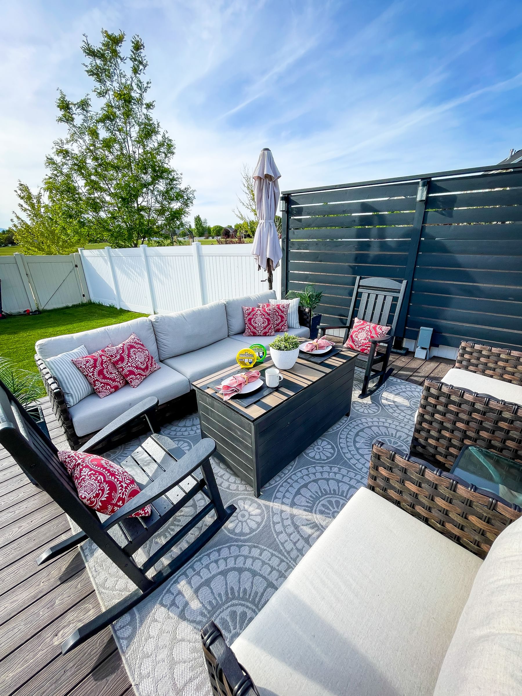 Stylish outdoor patio with a cozy gray sectional, vibrant red pillows, and a dark wood table set for snacks. Surrounded by a green lawn and white fence.