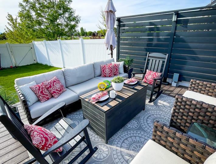 Stylish outdoor patio with a cozy gray sectional, vibrant red pillows, and a dark wood table set for snacks. Surrounded by a green lawn and white fence.