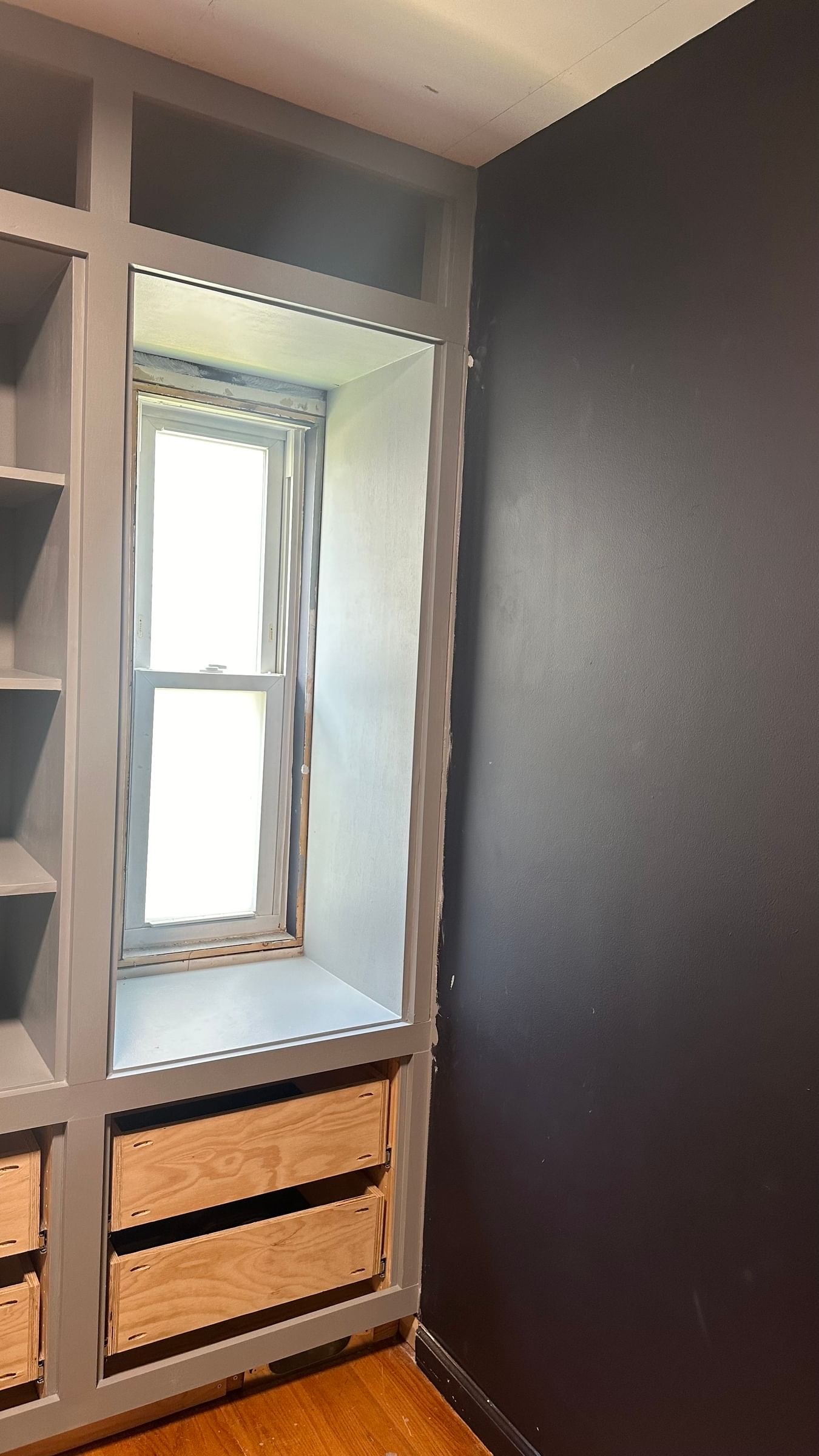 Built-in shelving unit with light gray frame, open wood drawers, and a recessed window with natural light. Adjacent wall painted dark gray. Cozy, modern feel.