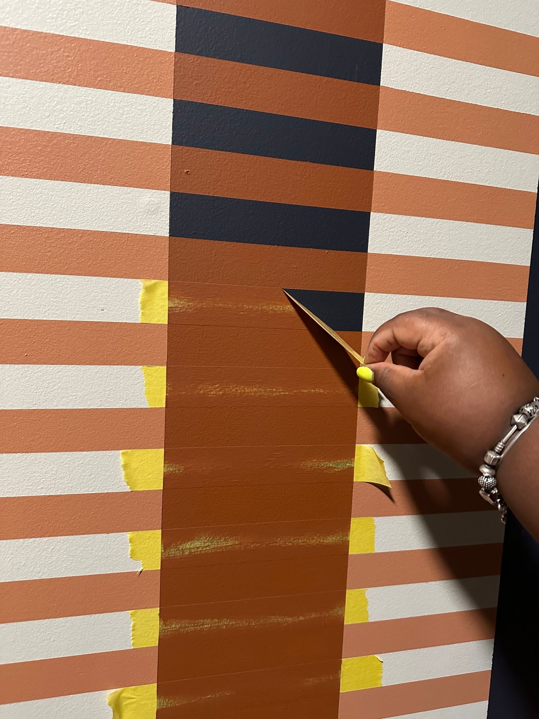 A hand with yellow-painted nails peels tape from a painted wall with horizontal stripes in shades of orange, navy, and white, conveying a creative DIY vibe.