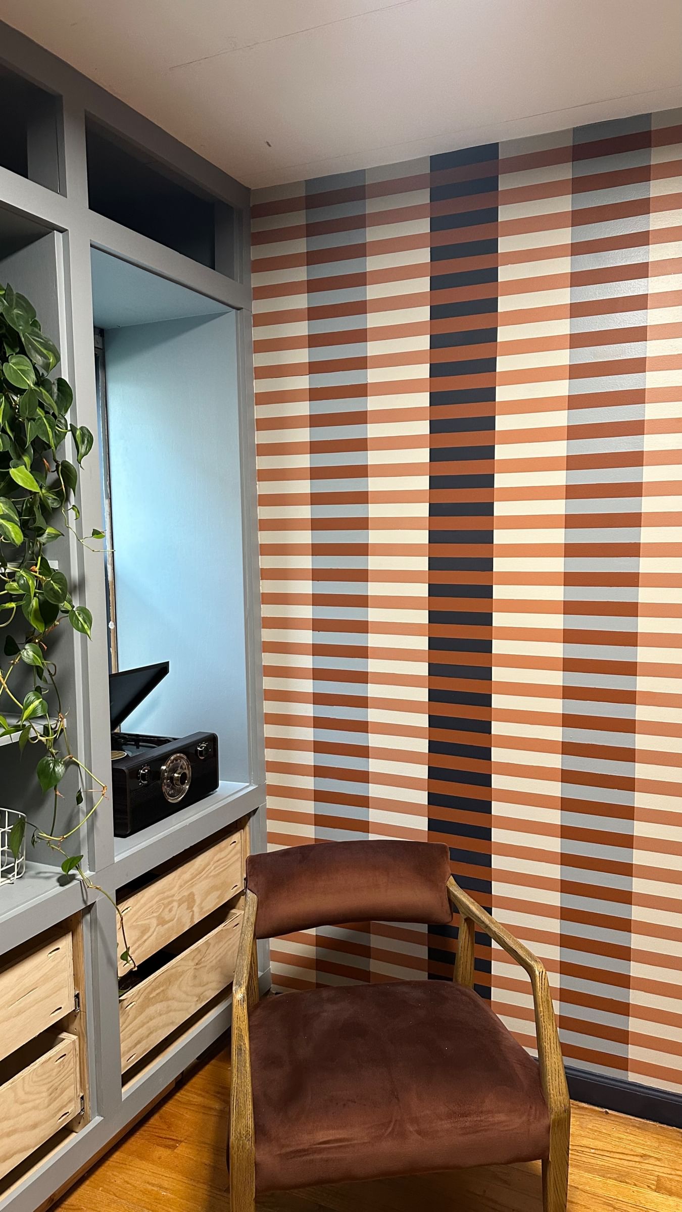 Cozy room with a striped accent wall in brown hues, modern shelving with plants and a record player. Brown velvet chair adds warmth to the hardwood floor.