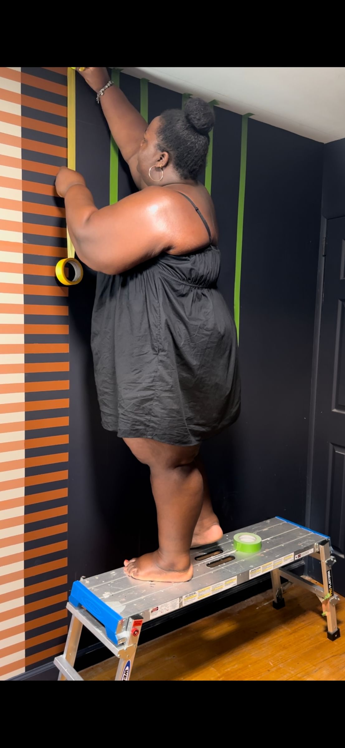 A person stands on a stepladder, applying green tape to a dark wall. Striped orange and black paneling is visible. The scene conveys focus and creativity.
