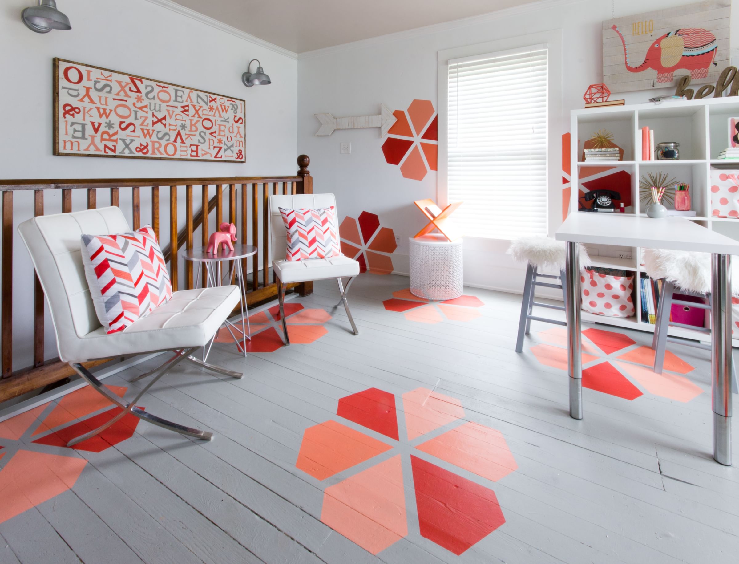 Bright, modern room with white chairs, red geometric cushions, colorful wall art, and hexagonal floor patterns. Shelving unit holds books and decor. Cozy and playful vibe.
