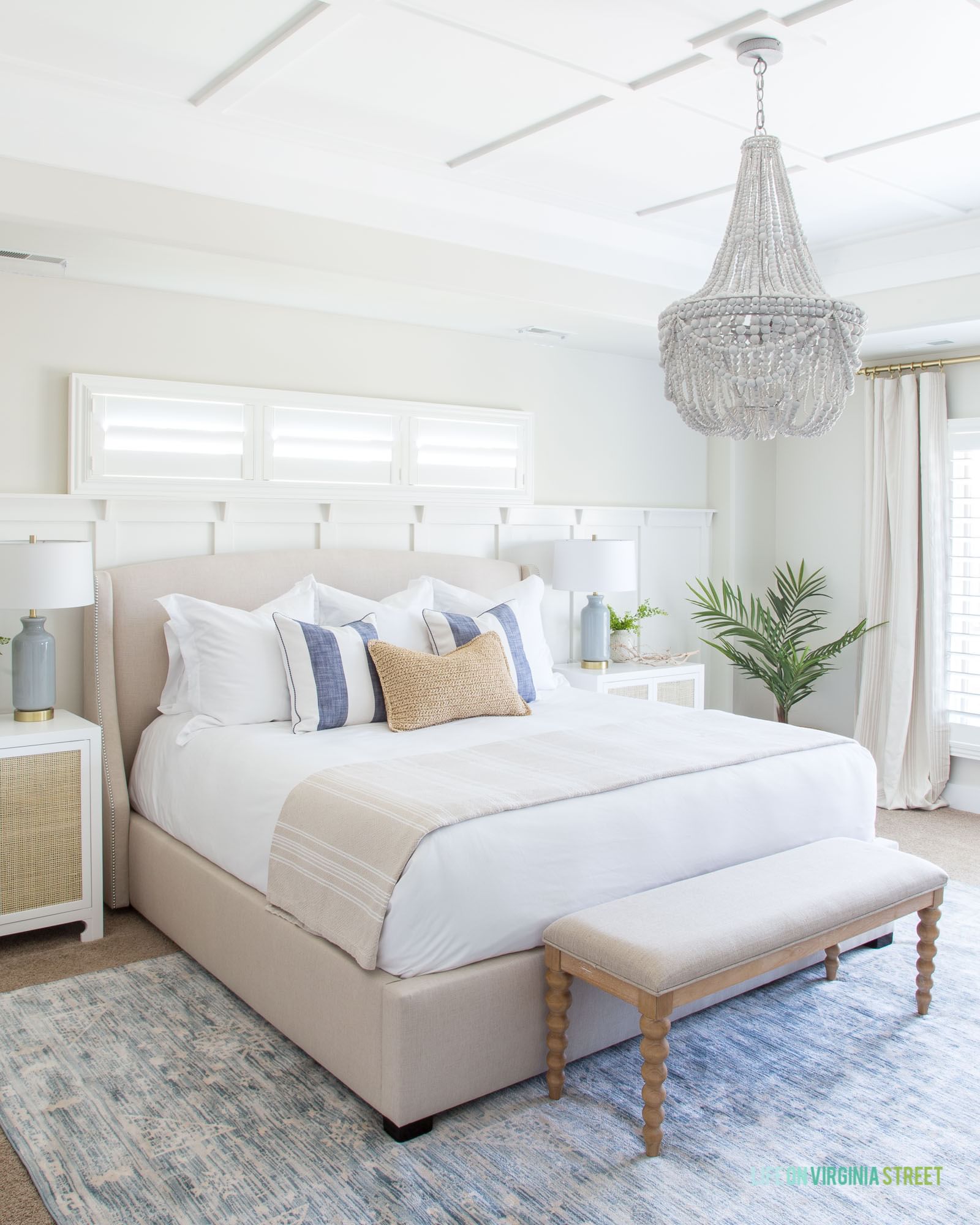 Elegant bedroom with neutral tones; features a plush bed with blue and beige pillows, a bench at the foot, white nightstands, and a beaded chandelier.