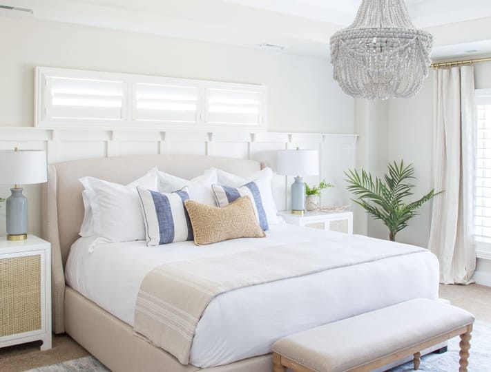 Elegant bedroom with neutral tones; features a plush bed with blue and beige pillows, a bench at the foot, white nightstands, and a beaded chandelier.