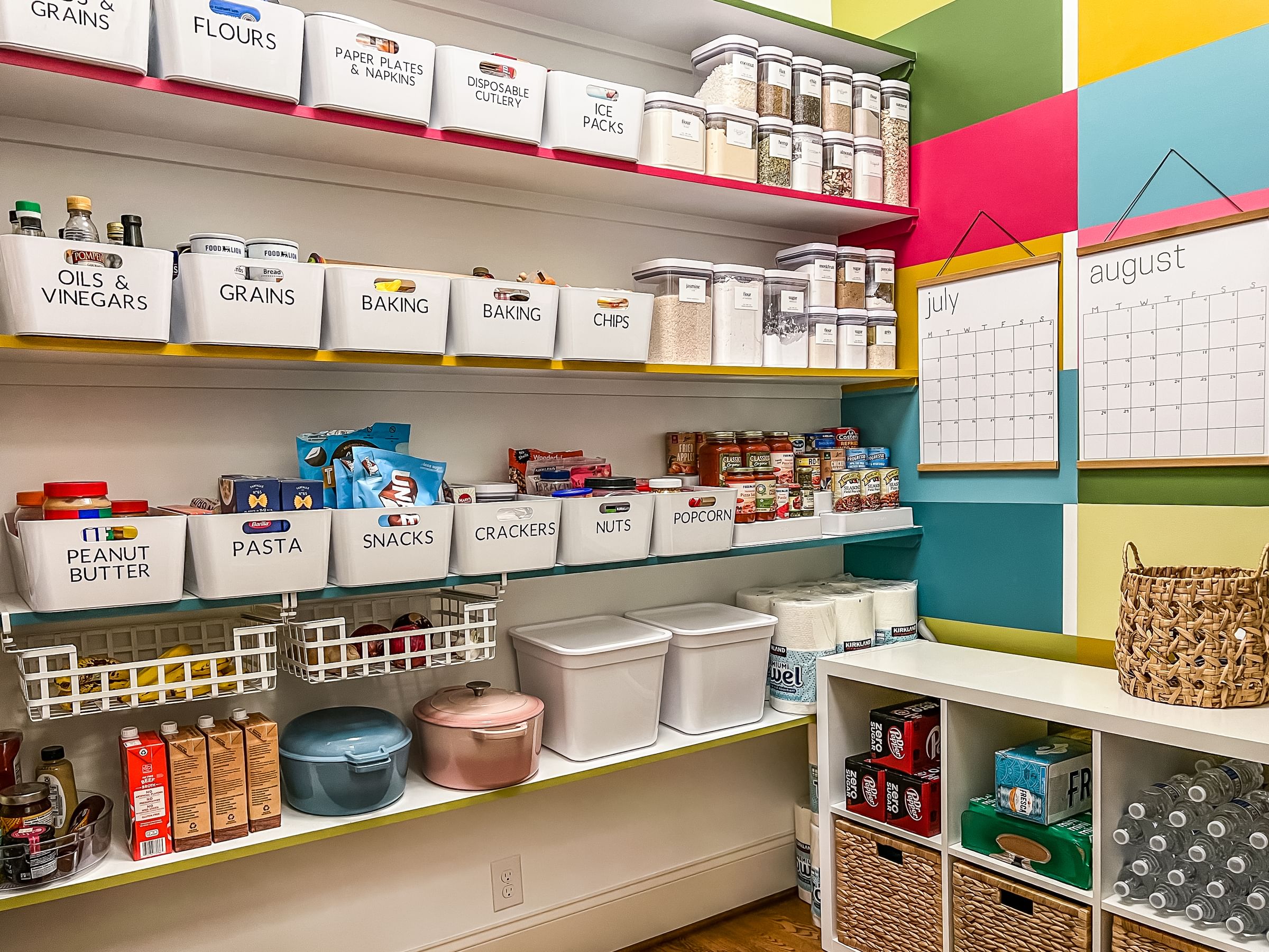 Bright, organized pantry with labeled bins for flours, snacks, and baking. Colorful wall with July and August calendars. Neat, cheerful atmosphere.