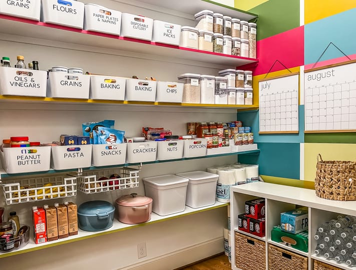 Bright, organized pantry with labeled bins for flours, snacks, and baking. Colorful wall with July and August calendars. Neat, cheerful atmosphere.