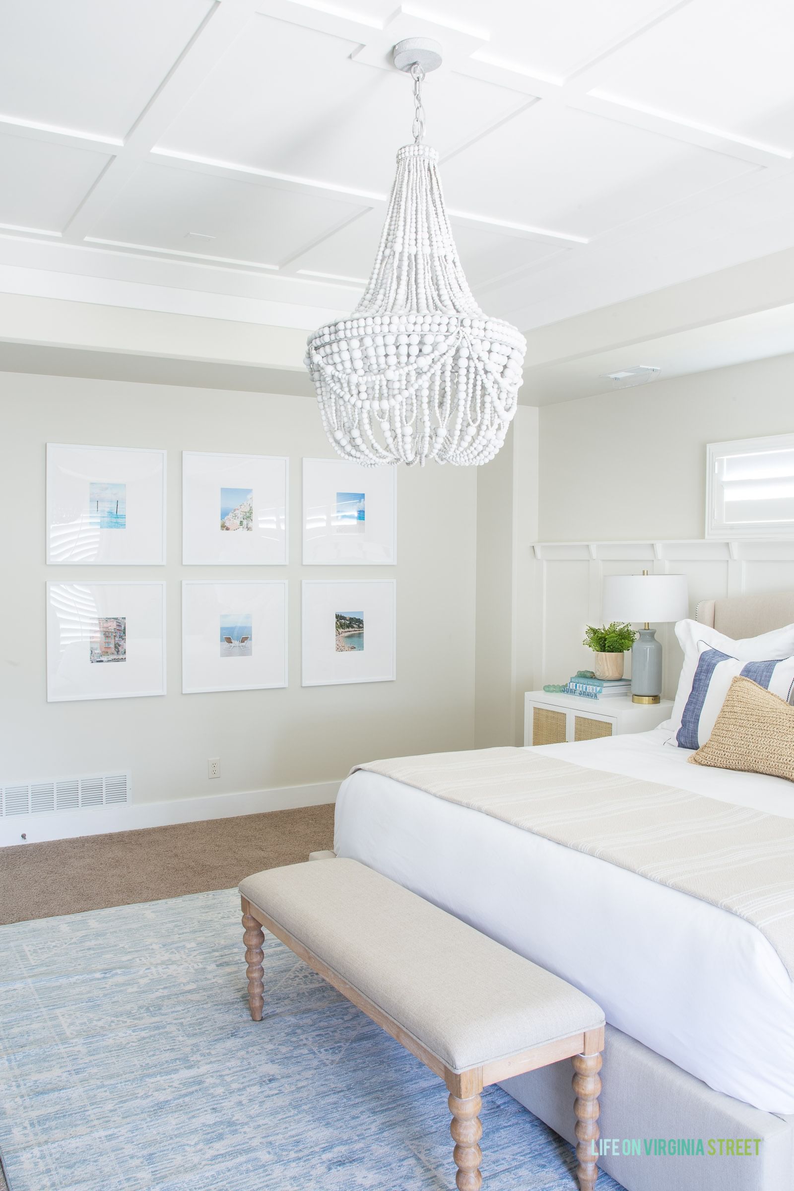 Bright, serene bedroom with a beige bed, cushioned bench, blue rug, and white chandelier. Art frames adorn the wall; decor has a coastal theme.