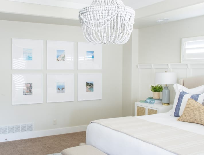 Bright, serene bedroom with a beige bed, cushioned bench, blue rug, and white chandelier. Art frames adorn the wall; decor has a coastal theme.