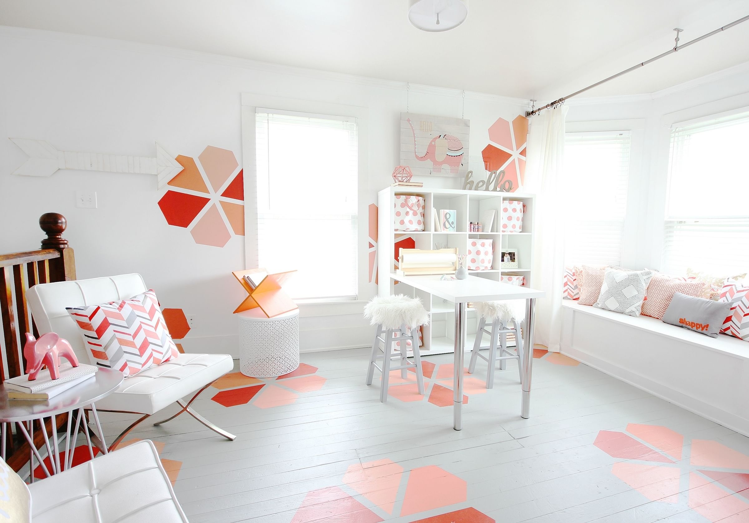Bright, modern room with a white and pink color scheme. Features geometric wall and floor patterns, a white table, and cozy seating with decorative pillows.