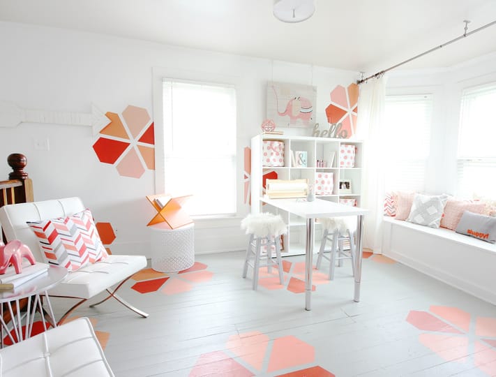 Bright, modern room with a white and pink color scheme. Features geometric wall and floor patterns, a white table, and cozy seating with decorative pillows.