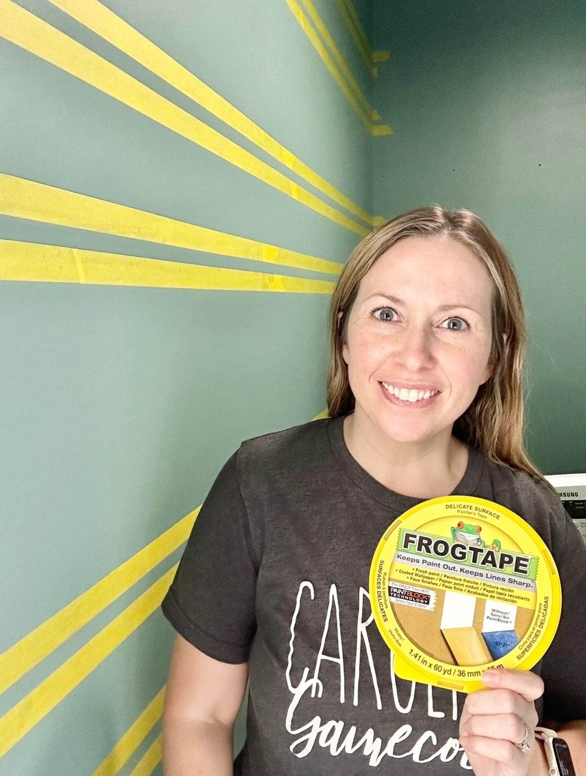 Katie Sharpe holding a Yellow FrogTape Delicate Surface Painter's Tape Cannister. Behind her is a blue/green wall will horizontal lines of the yellow tape