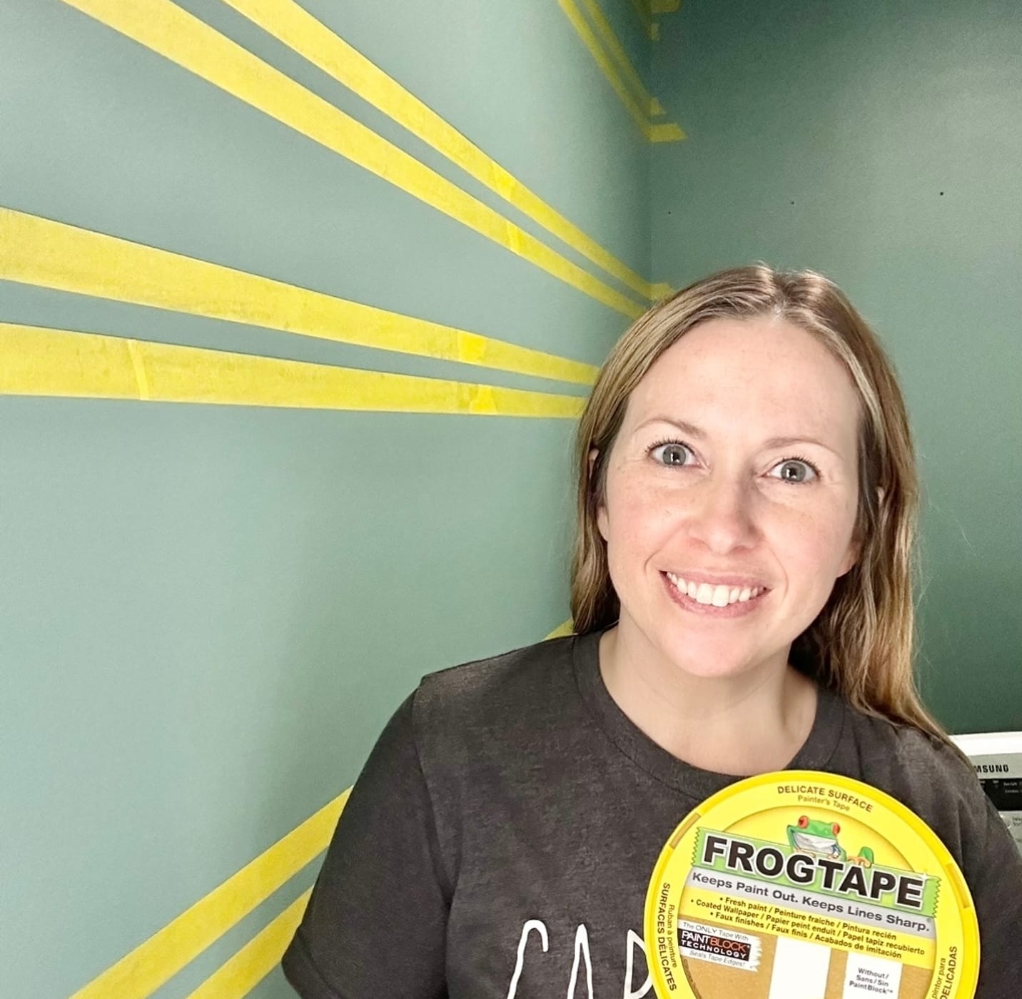 Katie Sharpe holding a Yellow FrogTape Delicate Surface Painter's Tape Cannister. Behind her is a blue/green wall will horizontal lines of the yellow tape