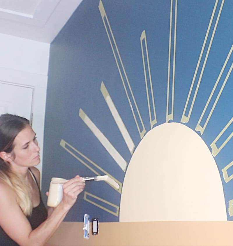 Woman painting the yellow sun rays on a sunset mural. She is using FrogTape for straight sunrays.