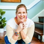 A cheerful person with blonde hair smiles brightly while leaning on a stair railing. They wear a striped shirt, colorful earrings, and bracelets. A potted plant is in the background.